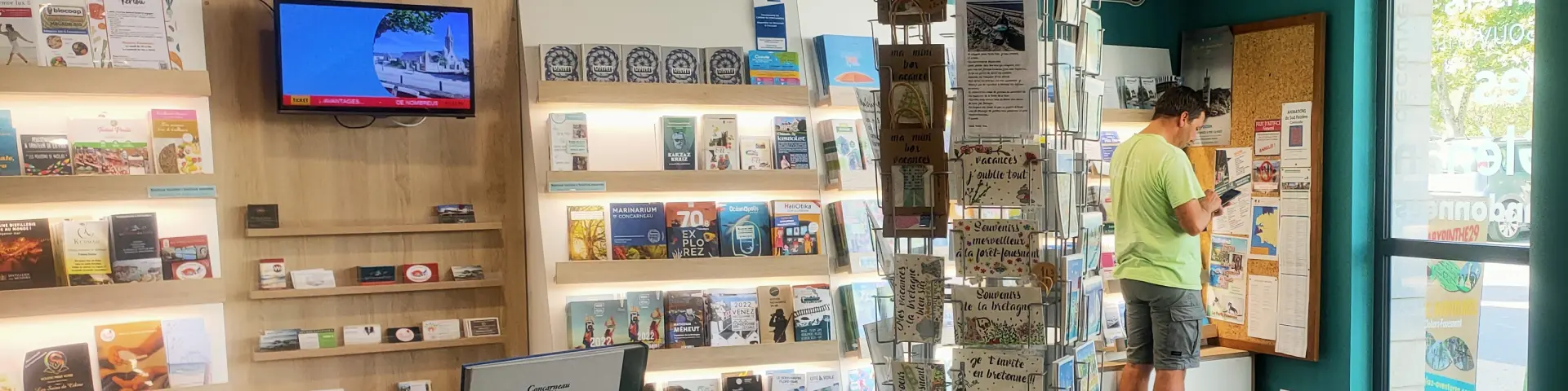 Un photo montrant l’intérieur d’un office de tourisme. La pièce a des murs turquoise, un comptoir en bois clair en forme courbe, un présentoir à cartes postales au milieu, des étagères remplies de brochures et livres, et un écran TV accroché au mur. Deux grands piliers cylindriques turquoise se trouvent près de l’entrée vitrée à droite. Une personne est debout près d’un panneau d’affichage au fond; une autre silhouette est visible vers la porte.