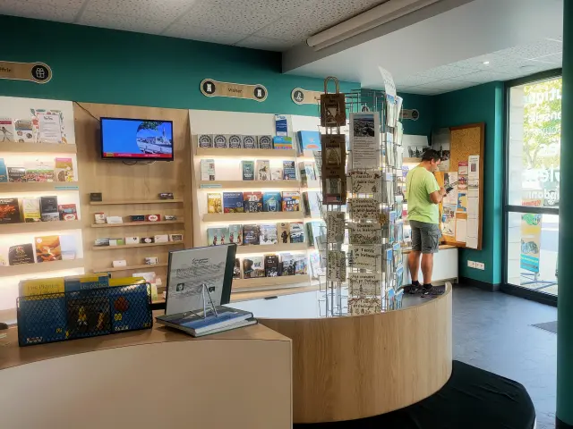 Een foto van het interieur van een VVV-kantoor. De kamer heeft turquoise muren, een gebogen toonbank van licht hout, een display met ansichtkaarten in het midden, planken vol brochures en boeken en een tv-scherm aan de muur. Bij de glazen ingang aan de rechterkant staan twee grote turquoise cilindervormige pilaren. Een persoon staat bij een prikbord aan de achterkant; een ander silhouet is zichtbaar in de richting van de deur.