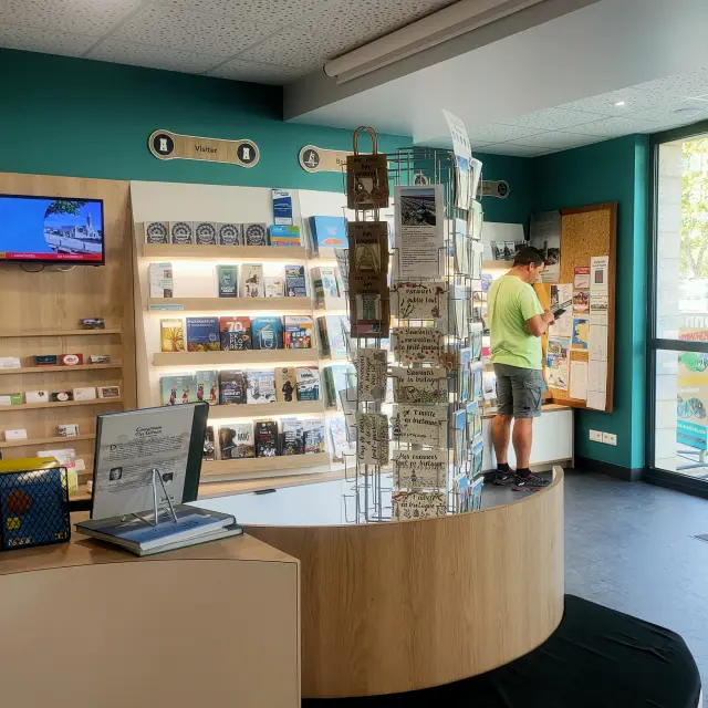 Un photo montrant l’intérieur d’un office de tourisme. La pièce a des murs turquoise, un comptoir en bois clair en forme courbe, un présentoir à cartes postales au milieu, des étagères remplies de brochures et livres, et un écran TV accroché au mur. Deux grands piliers cylindriques turquoise se trouvent près de l’entrée vitrée à droite. Une personne est debout près d’un panneau d’affichage au fond; une autre silhouette est visible vers la porte.