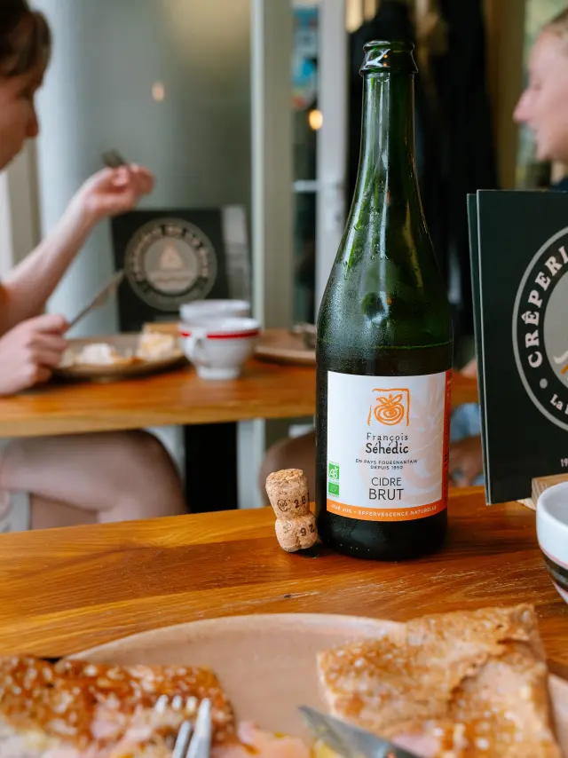 Foto van een fles cider op een houten tafel. Op het etiket staat La Forêt-Fouesnant en Cider BRUT. Er liggen twee kurken naast. Op de voorgrond een bord met een crêpe en wat bestek. Op de achtergrond zitten twee vage mensen aan tafel.