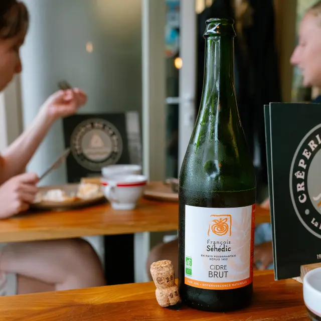 Foto einer Flasche Cidre, die auf einem Holztisch steht. Auf dem Etikett steht La Forêt-Fouesnant und Cider BRUT. Zwei Korken liegen daneben. Im Vordergrund stehen ein Teller mit einem Crêpe und Besteck. Im Hintergrund sitzen zwei verschwommene Personen am Tisch.