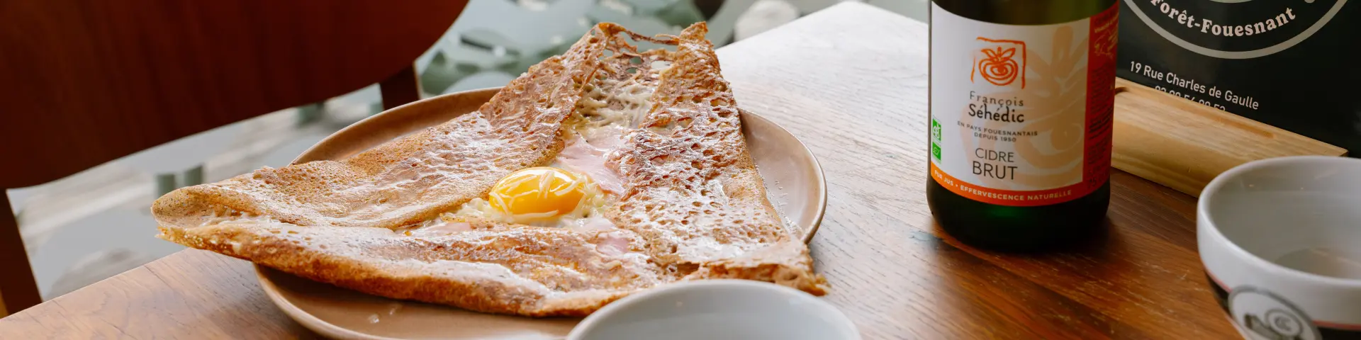 Deze foto toont een houten café/restauranttafel met een pannenkoek op een bord, een fles cider rechtopstaand, twee kommen/kopjes en bestek (vorken en messen) op een servet. Op de achtergrond van de foto suggereren gestapelde stoelen en tafels een terras. Een rond bord/voorwerp achter de fles toont een CRÊPERIE DE LA BAIE logo met een zeilontwerp; de tekst La Forêt-Fouesnant is zichtbaar op de cirkel. Op het etiket van de fles staat Cider BRUT.