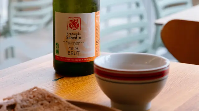 Une photo d'une bouteille en verre vert (type cidre) sur une table en bois, avec un bol blanc à liseré rouge à côté et une crêpe au premier plan sur une assiette. L’arrière-plan est une terrasse ou salle de café avec des chaises, floues.