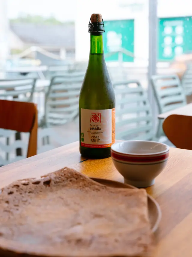 Une photo d'une bouteille en verre vert (type cidre) sur une table en bois, avec un bol blanc à liseré rouge à côté et une crêpe au premier plan sur une assiette. L’arrière-plan est une terrasse ou salle de café avec des chaises, floues.