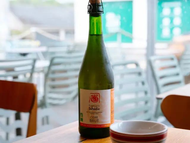 Ein Foto einer grünen Glasflasche (Typ Cidre) auf einem Holztisch, daneben eine weiße Schüssel mit rotem Rand und im Vordergrund ein Pfannkuchen auf einem Teller. Der Hintergrund ist eine Terrasse oder ein Café-Saal mit Stühlen, unscharf.