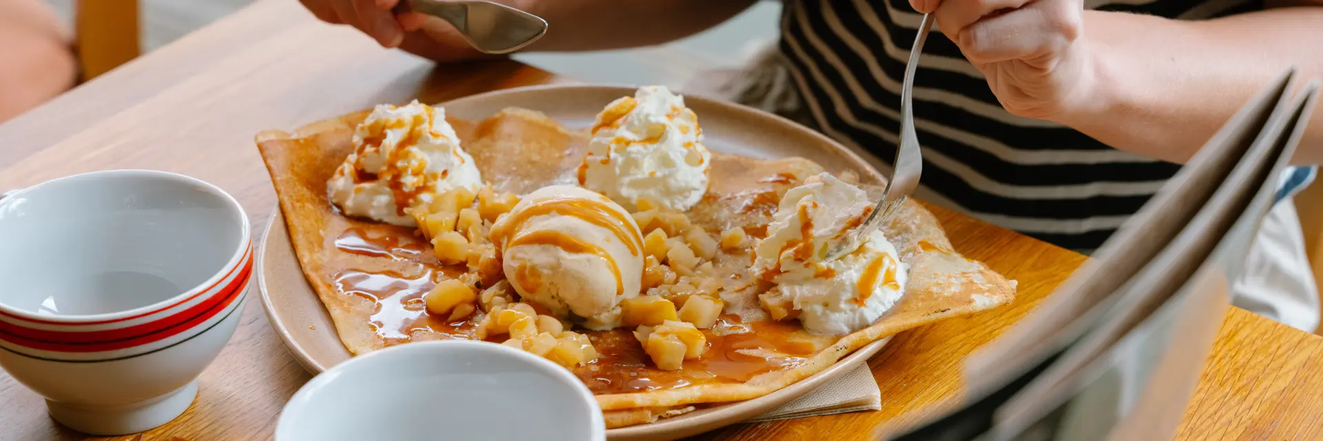 Foto einer Person mit einem schwarzen Oberteil mit weißen Streifen, die an einem Holztisch sitzt und einen großen Crêpe von einem Teller isst. Der Pfannkuchen ist mit mehreren Kugeln Eis oder Sahne, Stücken (Typ Trockenfrüchte/Haselnüsse) und einem Karamellüberzug belegt. Die Person hält einen Löffel und eine Gabel. Zwei weiße und rote Bolées stehen auf dem Tisch, und im Vordergrund ist ein kleines Stück Crêpe zu sehen.