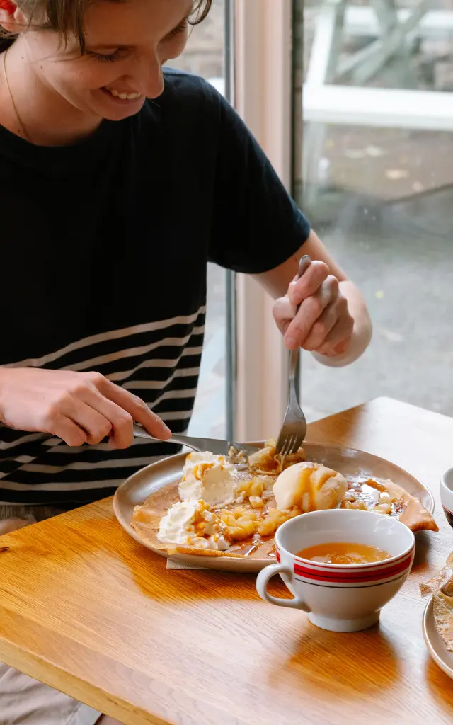 Das Foto zeigt zwei Frauen, die in einer Crêperie an einem Holztisch sitzen und essen. Vor der Frau rechts steht ein großer Crêpe auf einem Teller. Vor der Frau zu ihrer Linken befindet sich auf einem Teller eine Schüssel mit einem Ei. Auf dem Tisch stehen zwei Schalen mit Cidre. Ein kleines Schild mit der Aufschrift Crêperie de la Baie ist auf dem Tisch neben dem Fenster zu sehen.