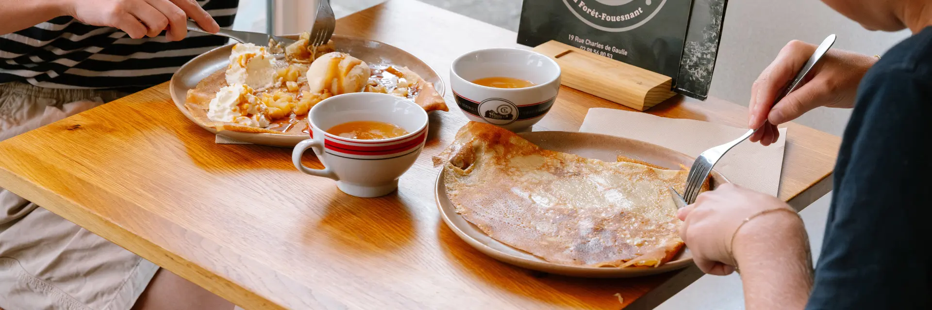 Das Foto zeigt zwei Frauen, die in einer Crêperie an einem Holztisch sitzen und essen. Vor der Frau rechts steht ein großer Crêpe auf einem Teller. Vor der Frau zu ihrer Linken befindet sich auf einem Teller eine Schüssel mit einem Ei. Auf dem Tisch stehen zwei Schalen mit Cidre. Ein kleines Schild mit der Aufschrift Crêperie de la Baie ist auf dem Tisch neben dem Fenster zu sehen.