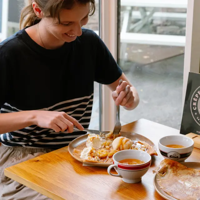 La photo montre deux femmes assises à une table en bois dans une crêperie, en train de manger. Une grande crêpe est posée sur une assiette devant la femme à droite. Devant la femme à gauche, une assiette contient un plat avec un œuf. Deux bolées de cidre sont sur la table. Un petit panneau “Crêperie de la Baie” est visible sur la table près de la fenêtre.