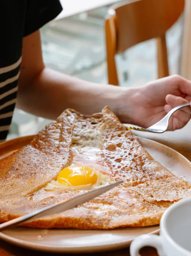 La photo montre une galette de sarrasin sur une assiette, garnie d’un œuf au centre (jaune visible). Une main tient une fourchette au-dessus de l’assiette et une autre main tient un couteau au bord de l’assiette. À droite de l’assiette, une bouteille est posée sur la table; l’étiquette indique “Cidre Brut” et “François Séhédic” . Deux bolées sont partiellement visibles au premier plan à droite.