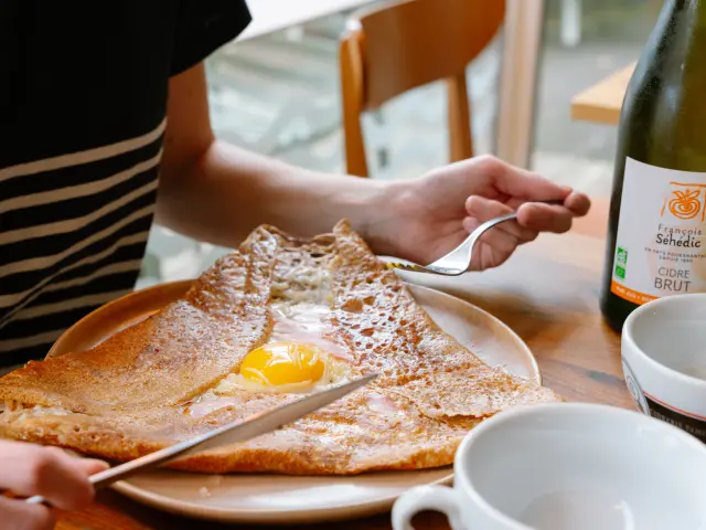 La photo montre une galette de sarrasin sur une assiette, garnie d’un œuf au centre (jaune visible). Une main tient une fourchette au-dessus de l’assiette et une autre main tient un couteau au bord de l’assiette. À droite de l’assiette, une bouteille est posée sur la table; l’étiquette indique “Cidre Brut” et “François Séhédic” . Deux bolées sont partiellement visibles au premier plan à droite.