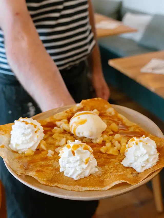 Une photo qui montre une assiette tenue au premier plan avec une grande crêpe garnie de plusieurs rosaces de crème chantilly et d’une boule de glace, avec un nappage (type caramel) et des morceaux de fruits à coque (type amandes/noisettes). En arrière-plan, une salle de restaurant est floue, avec des tables en bois et des banquettes.