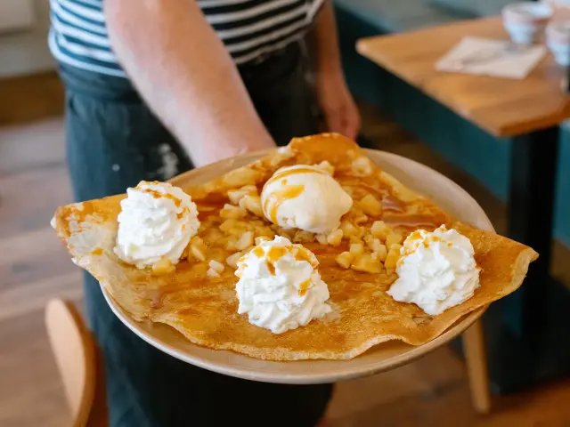 Une photo qui montre une assiette tenue au premier plan avec une grande crêpe garnie de plusieurs rosaces de crème chantilly et d’une boule de glace, avec un nappage (type caramel) et des morceaux de fruits à coque (type amandes/noisettes). En arrière-plan, une salle de restaurant est floue, avec des tables en bois et des banquettes.