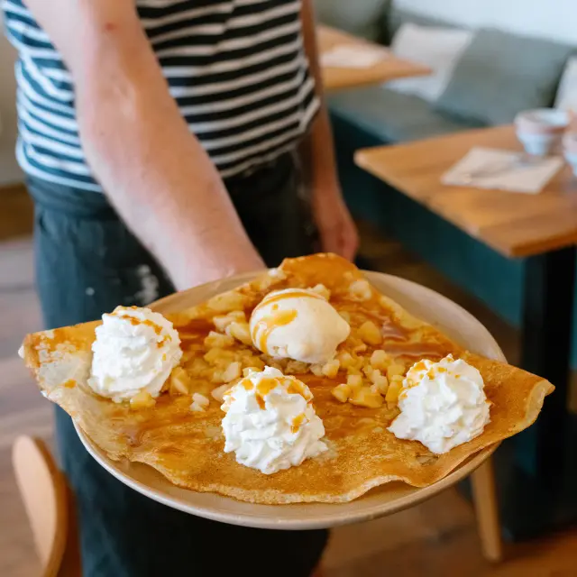 Ein Foto, das einen im Vordergrund gehaltenen Teller mit einem großen Crêpe zeigt, der mit mehreren Rosetten aus Schlagsahne und einer Kugel Eis belegt ist, mit einem Topping (Typ Karamell) und Nussstücken (Typ Mandeln/Haselnüsse). Im Hintergrund ist verschwommen ein Gastraum mit Holztischen und Sitzbänken zu sehen.