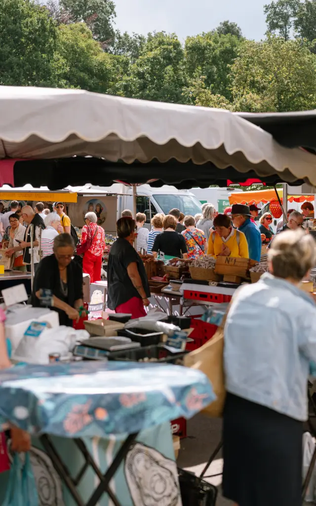 Photo d’un marché en plein air. La scène montre une foule autour de stands sous des auvents beige et noir; plusieurs personnes sont au premier plan près de tables hautes, et des arbres sont visibles à l’arrière-plan.