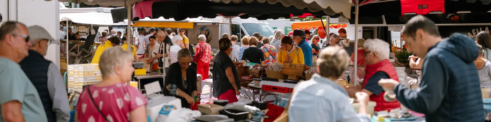 Photo d’un marché en plein air. La scène montre une foule autour de stands sous des auvents beige et noir; plusieurs personnes sont au premier plan près de tables hautes, et des arbres sont visibles à l’arrière-plan.