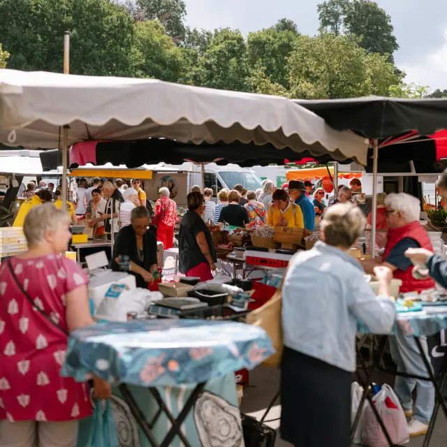 Photo d’un marché en plein air. La scène montre une foule autour de stands sous des auvents beige et noir; plusieurs personnes sont au premier plan près de tables hautes, et des arbres sont visibles à l’arrière-plan.