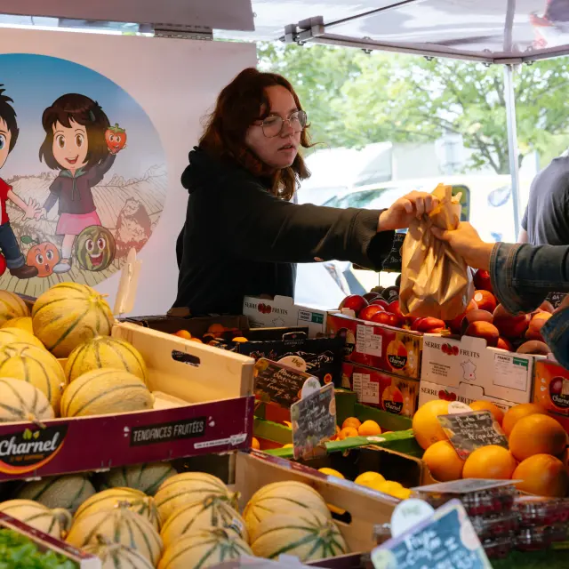 La photo montre un étal de marché avec des cagettes de melons (jaune clair avec des rayures vertes), des tomates et d’autres fruits (oranges/jaunes). Deux femmes sont au centre: l’une (cheveux bruns, lunettes, vêtement sombre) tend un sachet en papier; l’autre (cheveux blonds, veste en jean) le reçoit. À l’arrière de l’étal, un grand panneau illustré avec des personnages dessinés (style cartoon) dans un cercle.
