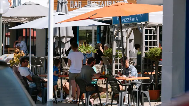 La photo montre une terrasse de restaurant en extérieur, avec plusieurs tables et chaises occupées par des personnes. De grands parasols blancs et un grand parasol orange couvrent les tables. À l’arrière, des enseignes sont visibles, dont “BAR” et “PIZZÉRIA”.