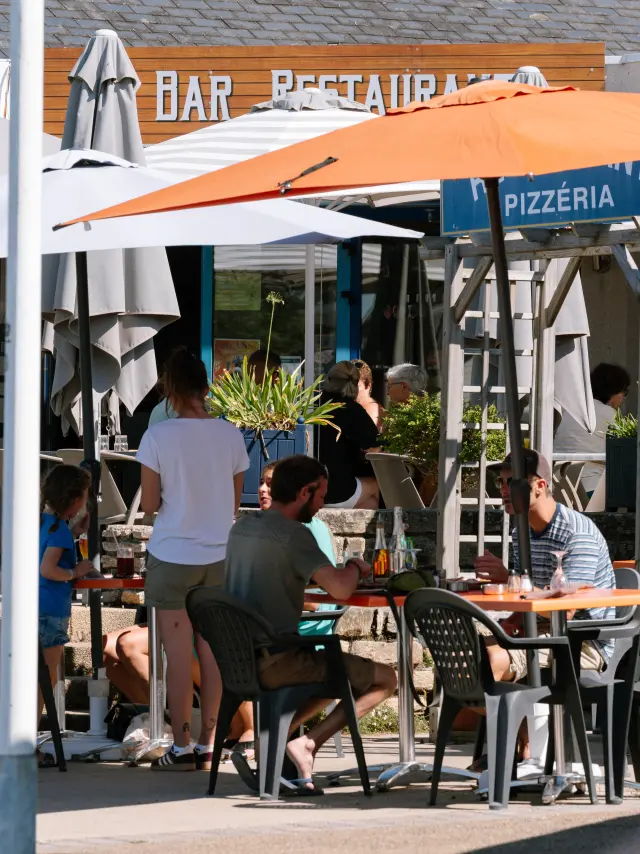 La photo montre une terrasse de restaurant en extérieur, avec plusieurs tables et chaises occupées par des personnes. De grands parasols blancs et un grand parasol orange couvrent les tables. À l’arrière, des enseignes sont visibles, dont “BAR” et “PIZZÉRIA”.