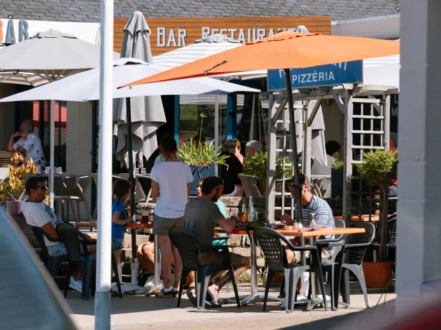 La photo montre une terrasse de restaurant en extérieur, avec plusieurs tables et chaises occupées par des personnes. De grands parasols blancs et un grand parasol orange couvrent les tables. À l’arrière, des enseignes sont visibles, dont “BAR” et “PIZZÉRIA”.