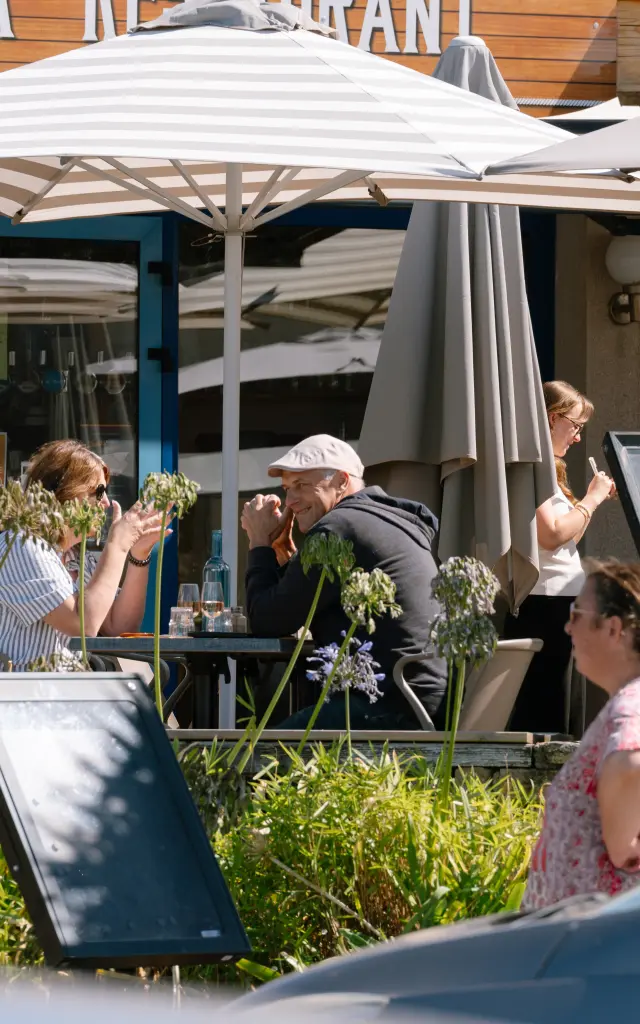 Une photo montrant une terrasse de restaurant. Plusieurs personnes sont assises et discutent à des tables sous de grands parasols blancs; d’autres personnes passent devant. Au premier plan, une voiture sombre occupe le bas droit de l’image.
