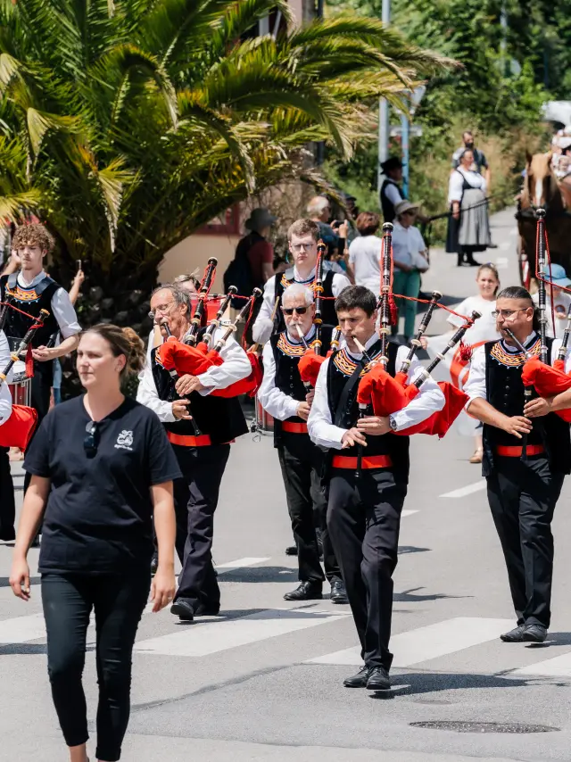 Une photo d’un défilé : un groupe de musiciens en tenue bretonne (gilets noirs, chemises blanches, éléments rouges) marche dans une rue en jouant de la cornemuse; un joueur de caisse claire est visible. Une foule suit derrière; un grand palmier est sur la gauche de la scène.