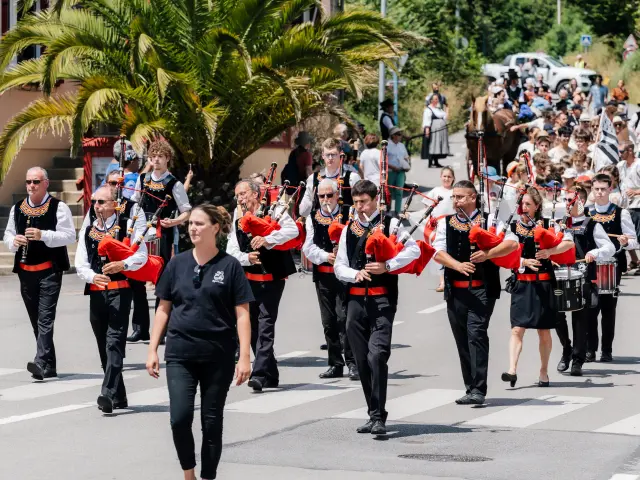Une photo d’un défilé : un groupe de musiciens en tenue bretonne (gilets noirs, chemises blanches, éléments rouges) marche dans une rue en jouant de la cornemuse; un joueur de caisse claire est visible. Une foule suit derrière; un grand palmier est sur la gauche de la scène.