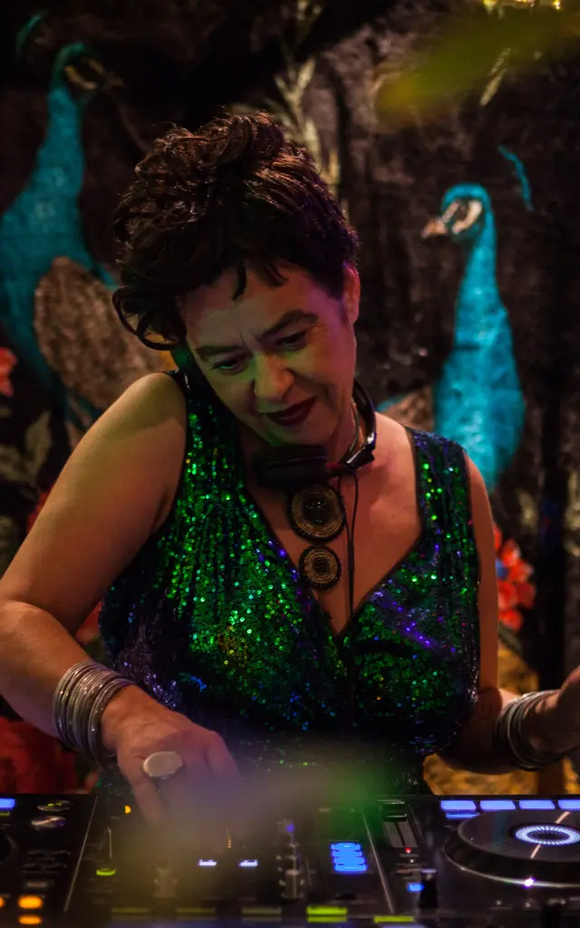 La photo montre une personne en train de mixer sur une table de DJ (deux platines avec anneaux lumineux). La personne porte une robe ou un haut vert scintillant, des bracelets, et un grand collier. L’arrière-plan est une tenture sombre avec des motifs floraux rouges et deux oiseaux de type paon bleus, avec une guirlande lumineuse au-dessus.