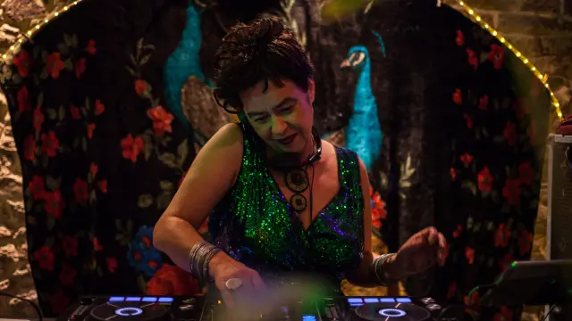 De foto toont een persoon die aan het mixen is op een DJ-tafel (twee draaitafels met verlichte ringen). De persoon draagt een sprankelende groene jurk of top, armbanden en een grote ketting. De achtergrond is een donkere ophanging met rode bloemmotieven en twee blauwe pauwenvogels, met een lichtsnoer erboven.