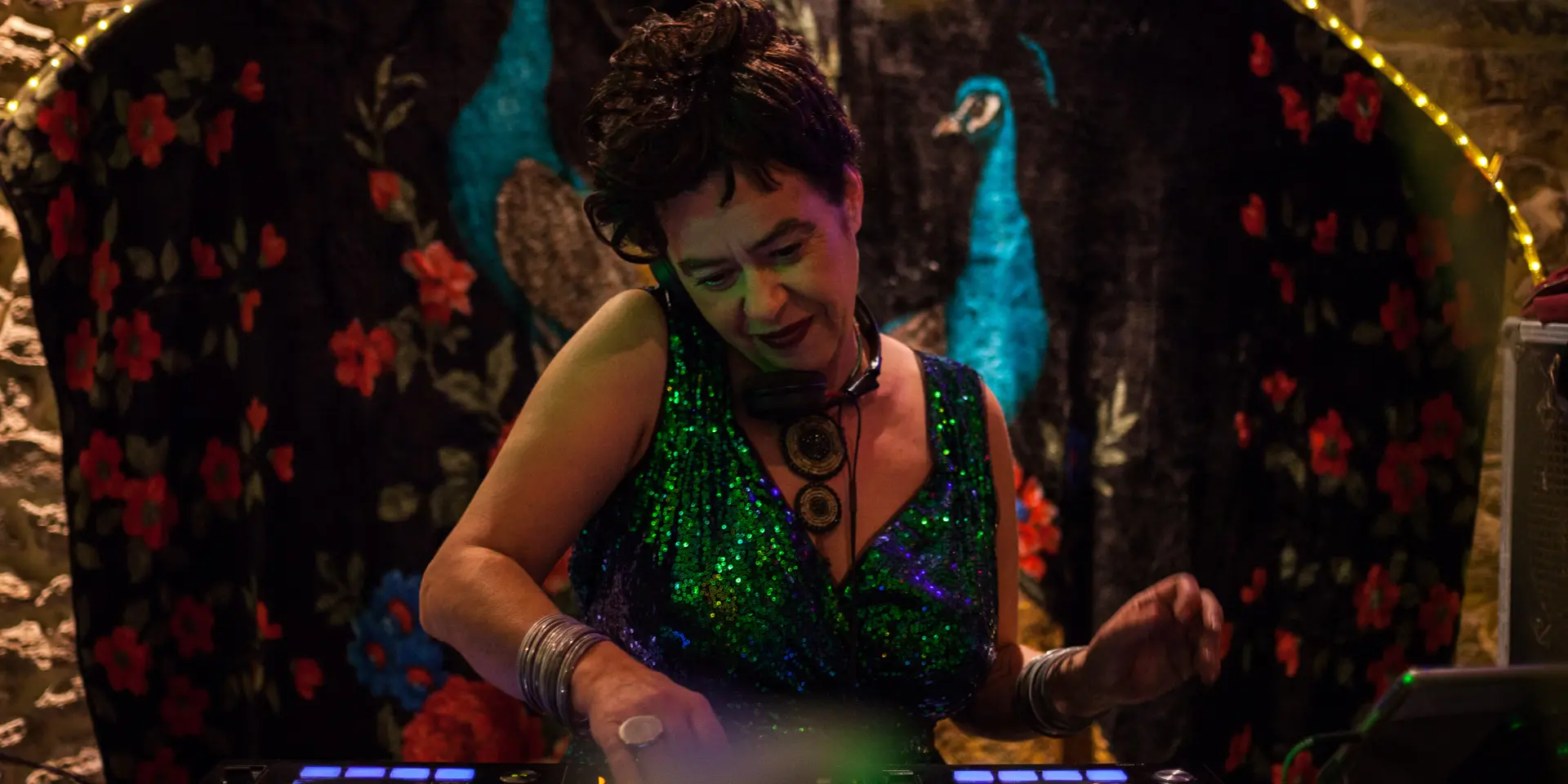 De foto toont een persoon die aan het mixen is op een DJ-tafel (twee draaitafels met verlichte ringen). De persoon draagt een sprankelende groene jurk of top, armbanden en een grote ketting. De achtergrond is een donkere ophanging met rode bloemmotieven en twee blauwe pauwenvogels, met een lichtsnoer erboven.
