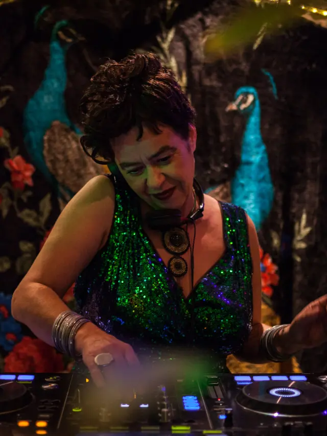 La photo montre une personne en train de mixer sur une table de DJ (deux platines avec anneaux lumineux). La personne porte une robe ou un haut vert scintillant, des bracelets, et un grand collier. L’arrière-plan est une tenture sombre avec des motifs floraux rouges et deux oiseaux de type paon bleus, avec une guirlande lumineuse au-dessus.