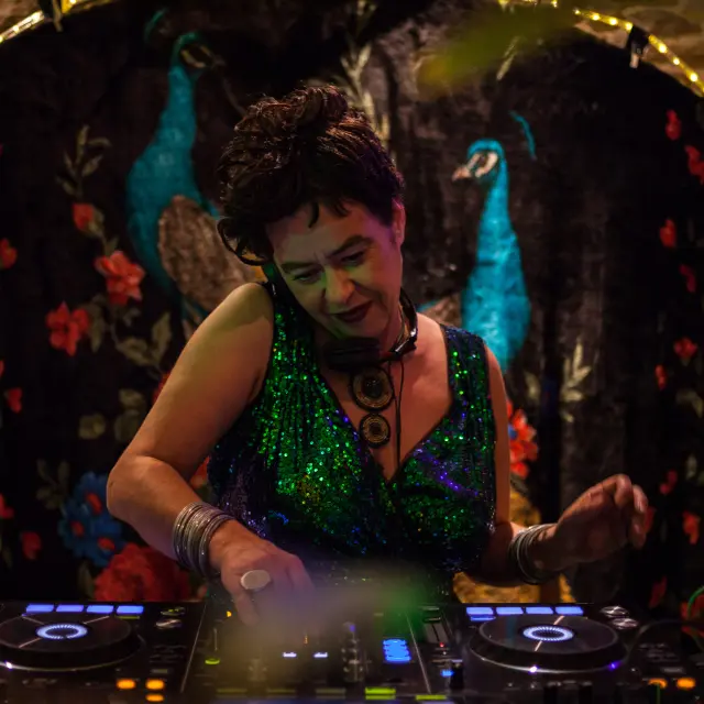 De foto toont een persoon die aan het mixen is op een DJ-tafel (twee draaitafels met verlichte ringen). De persoon draagt een sprankelende groene jurk of top, armbanden en een grote ketting. De achtergrond is een donkere ophanging met rode bloemmotieven en twee blauwe pauwenvogels, met een lichtsnoer erboven.