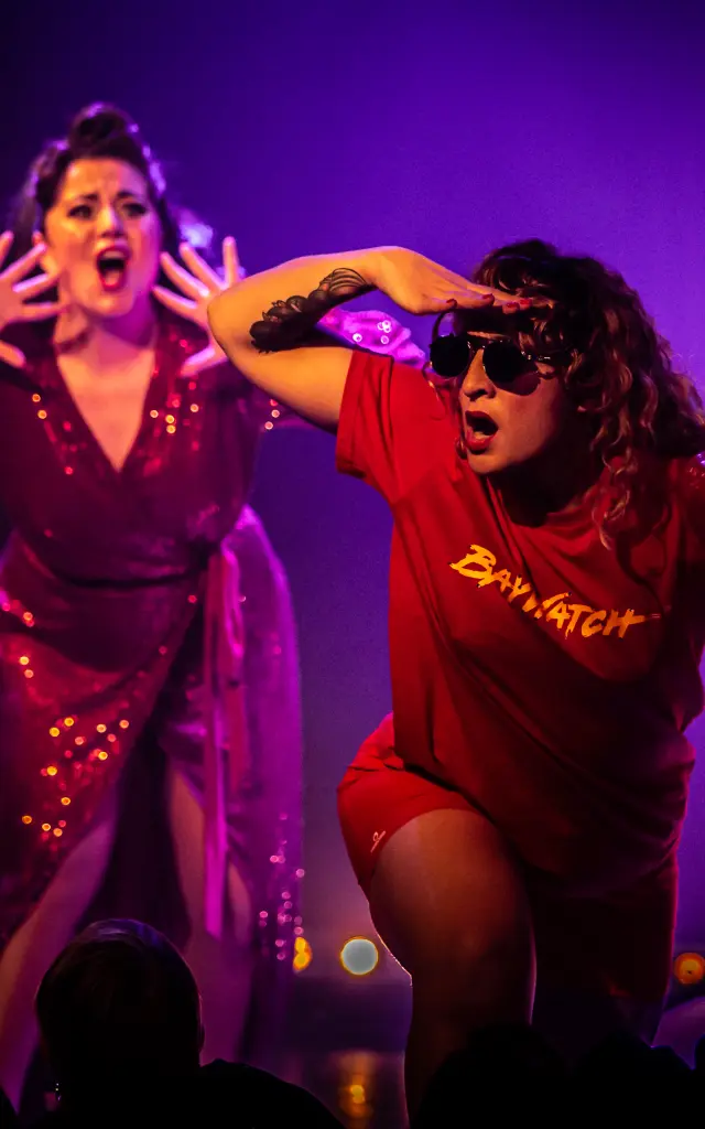 Photo qui montre deux personnes sur scène sous des éclairages violets et roses, en tenue de spectacle, avec des silhouettes de public au bas de l’image.