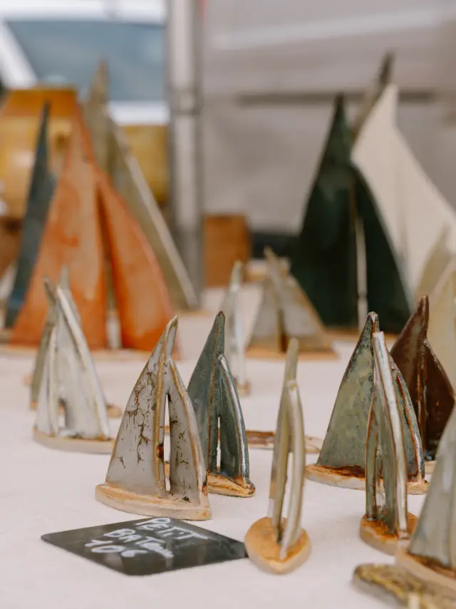 Une photo qui montre de nombreux petits bateaux en céramique posés sur une table claire, avec des pièces de différentes couleurs (gris, brun, vert, orangé) et des vases flous en arrière-plan.