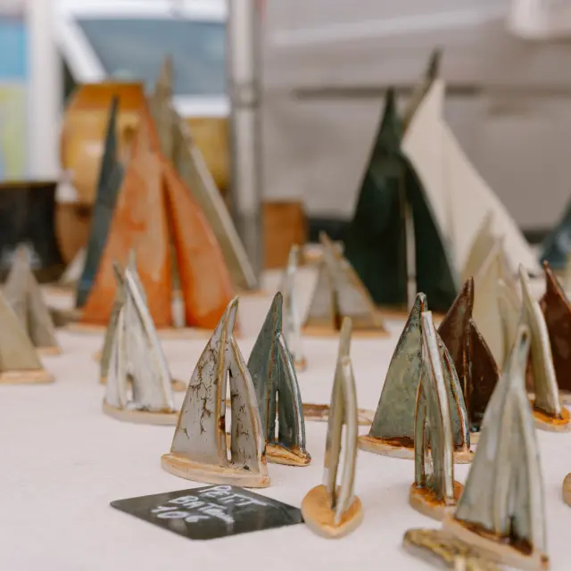 Une photo qui montre de nombreux petits bateaux en céramique posés sur une table claire, avec des pièces de différentes couleurs (gris, brun, vert, orangé) et des vases flous en arrière-plan.