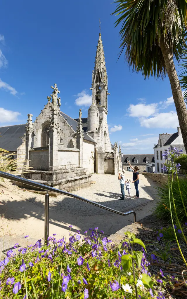 La photo montre une église en pierre claire avec une flèche très haute, sous un ciel bleu. Un chemin mène vers l’église, avec des fleurs violettes au premier plan et un palmier sur la droite. Trois personnes admirent l'église.