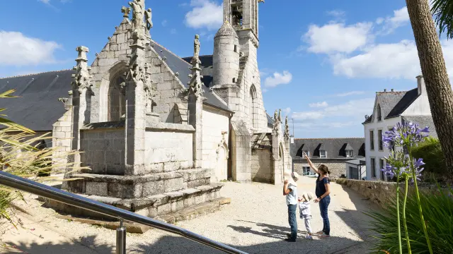 La photo montre une église en pierre claire avec une flèche très haute, sous un ciel bleu. Un chemin mène vers l’église, avec des fleurs violettes au premier plan et un palmier sur la droite. Trois personnes admirent l'église.