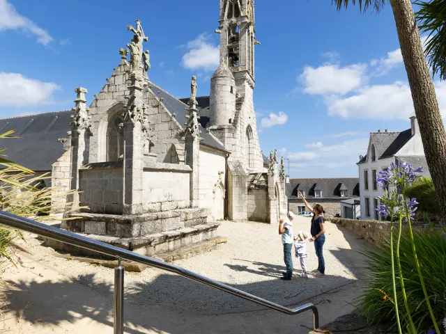 La photo montre une église en pierre claire avec une flèche très haute, sous un ciel bleu. Un chemin mène vers l’église, avec des fleurs violettes au premier plan et un palmier sur la droite. Trois personnes admirent l'église.