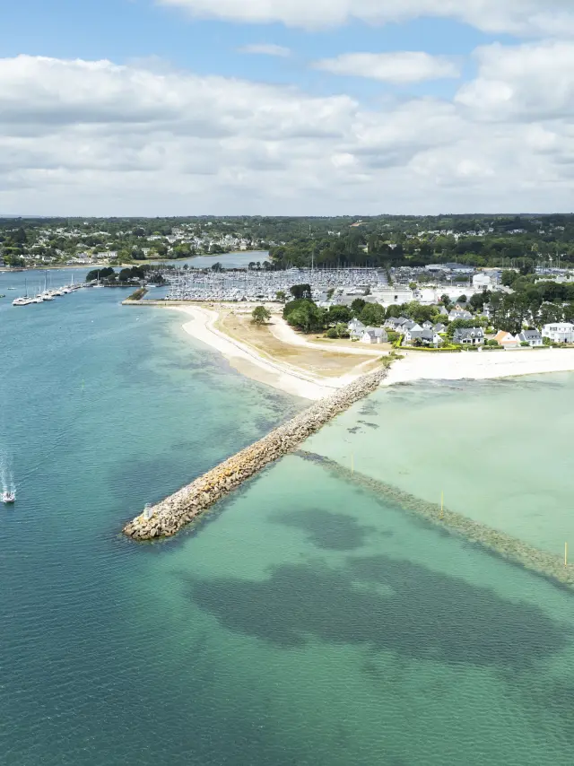 Une photo aérienne de littoral qui montre une plage de sable clair, une digue en pierres qui avance dans l’eau, et une mer allant du bleu foncé au turquoise/vert clair, avec plusieurs petits bateaux au mouillage. À l’arrière, des maisons, le port et de la végétation sous un ciel nuageux.