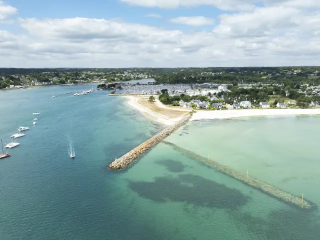Une photo aérienne de littoral qui montre une plage de sable clair, une digue en pierres qui avance dans l’eau, et une mer allant du bleu foncé au turquoise/vert clair, avec plusieurs petits bateaux au mouillage. À l’arrière, des maisons, le port et de la végétation sous un ciel nuageux.