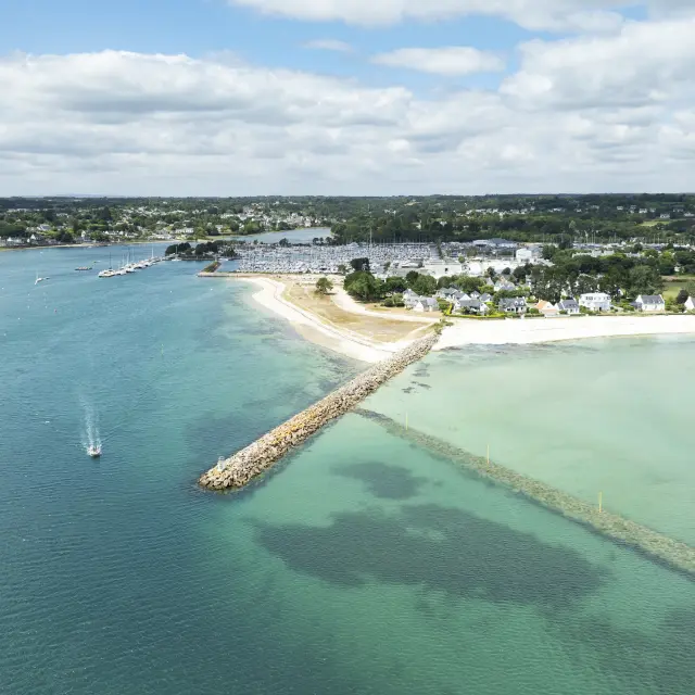 Une photo aérienne de littoral qui montre une plage de sable clair, une digue en pierres qui avance dans l’eau, et une mer allant du bleu foncé au turquoise/vert clair, avec plusieurs petits bateaux au mouillage. À l’arrière, des maisons, le port et de la végétation sous un ciel nuageux.