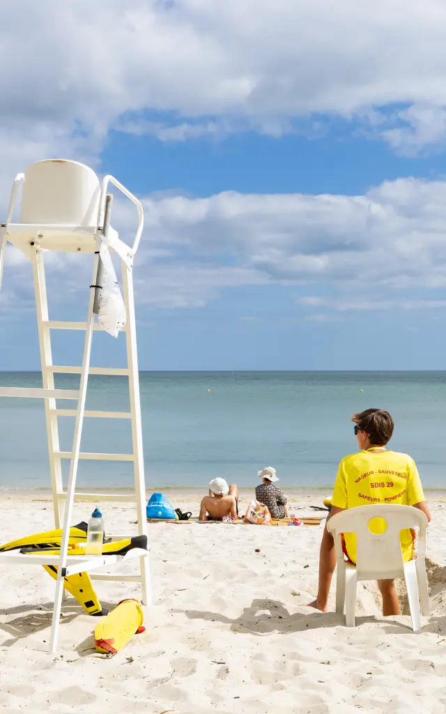 La photo montre une plage de sable clair, la mer calme à l’horizon et un ciel bleu avec de nombreux nuages. Une chaise haute de surveillance blanche est à gauche. Trois sauveteurs assis de dos portent des tee-shirts jaunes; quelques personnes sont allongées plus loin sur le sable. Du matériel jaune (type bouée/équipement de secours) est posé au sol près de la chaise.
