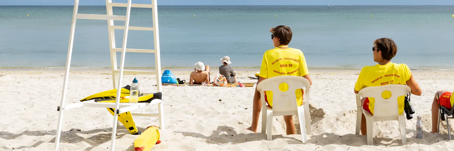 The photo shows a clear sandy beach, calm sea on the horizon and a blue sky with many clouds. A white lifeguard chair is on the left. Three lifeguards seated from behind are wearing yellow T-shirts; a few people are lying further back on the sand. Yellow equipment (buoy/rescue gear) is on the ground next to the chair.