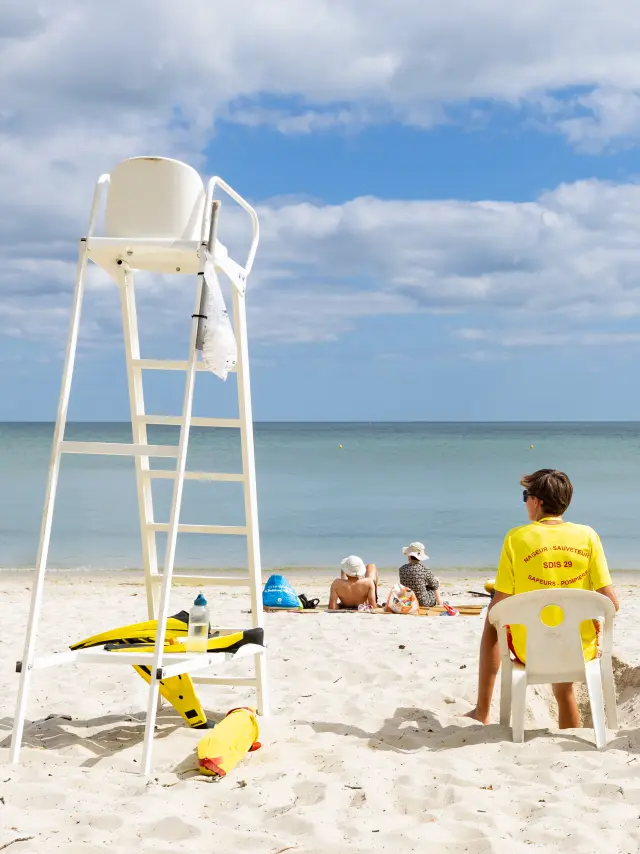 La photo montre une plage de sable clair, la mer calme à l’horizon et un ciel bleu avec de nombreux nuages. Une chaise haute de surveillance blanche est à gauche. Trois sauveteurs assis de dos portent des tee-shirts jaunes; quelques personnes sont allongées plus loin sur le sable. Du matériel jaune (type bouée/équipement de secours) est posé au sol près de la chaise.
