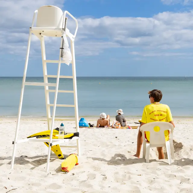 The photo shows a clear sandy beach, calm sea on the horizon and a blue sky with many clouds. A white lifeguard chair is on the left. Three lifeguards seated from behind are wearing yellow T-shirts; a few people are lying further back on the sand. Yellow equipment (buoy/rescue gear) is on the ground next to the chair.