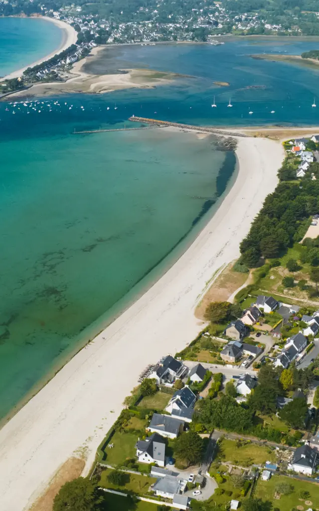 Une photo d'une vue aérienne d’un littoral : longue plage de sable blanc en courbe, eau turquoise et verte, un chenal avec bancs de sable, quelques bateaux au large, et des maisons regroupées près du rivage.