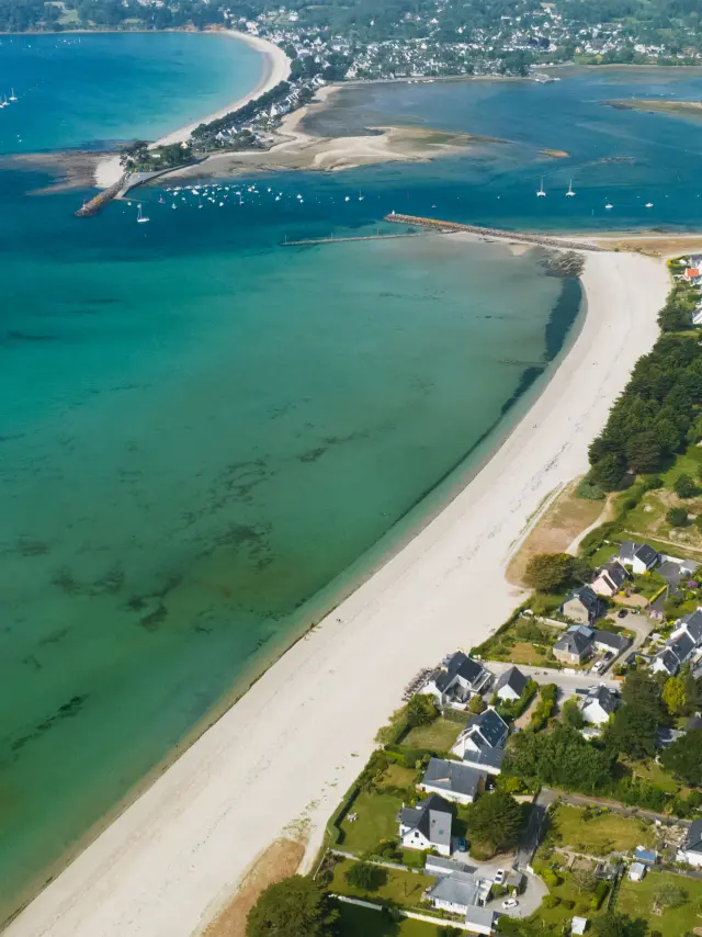 Une photo d'une vue aérienne d’un littoral : longue plage de sable blanc en courbe, eau turquoise et verte, un chenal avec bancs de sable, quelques bateaux au large, et des maisons regroupées près du rivage.