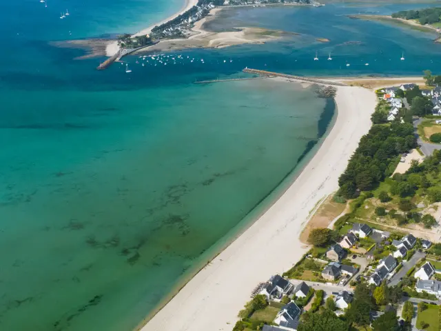 Une photo d'une vue aérienne d’un littoral : longue plage de sable blanc en courbe, eau turquoise et verte, un chenal avec bancs de sable, quelques bateaux au large, et des maisons regroupées près du rivage.
