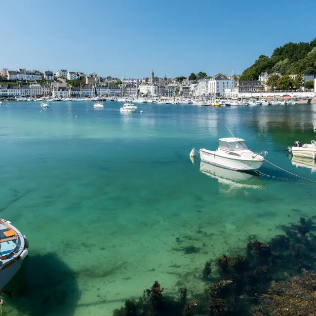 Une photo qui montre un port avec de l’eau turquoise très claire, plusieurs petits bateaux au mouillage, et une rangée de bâtiments blancs sur la rive sous un ciel bleu.